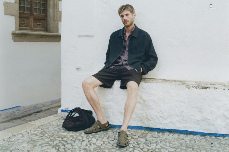 Blonde man in shorts and a jacket sits against a white wall in a cobblestone alley with a black bag and olive sandals.