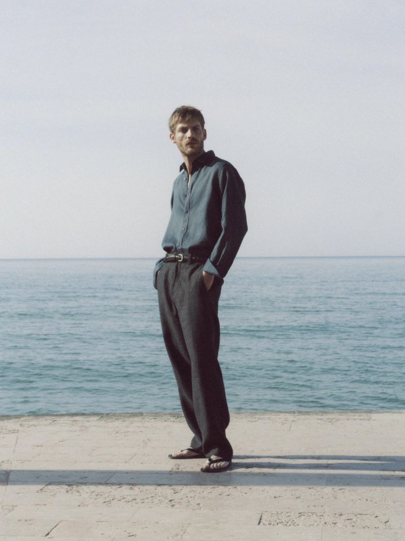 A stylish man in a blue shirt and grey trousers stands on a concrete surface near the ocean under a bright sky, fashion editorial.