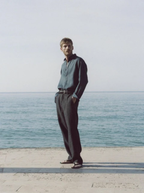 A stylish man in a blue shirt and grey trousers stands on a concrete surface near the ocean under a bright sky, fashion editorial.