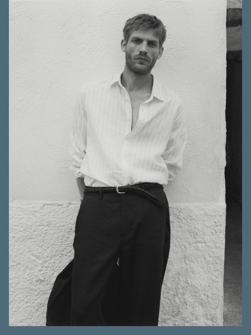 A man poses near a white wall, wearing an unbuttoned white shirt and black trousers, highlighted by black and white tones.
