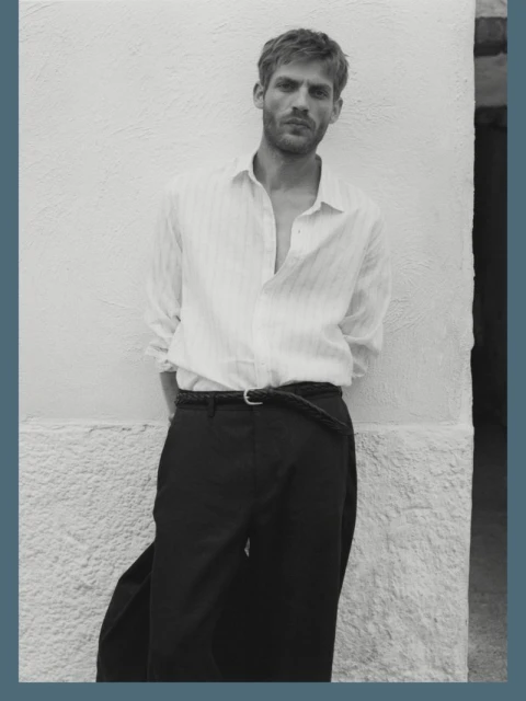 A man poses near a white wall, wearing an unbuttoned white shirt and black trousers, highlighted by black and white tones.