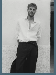 Black and white portrait of a man in a light collared shirt and dark trousers leaning against a textured wall.