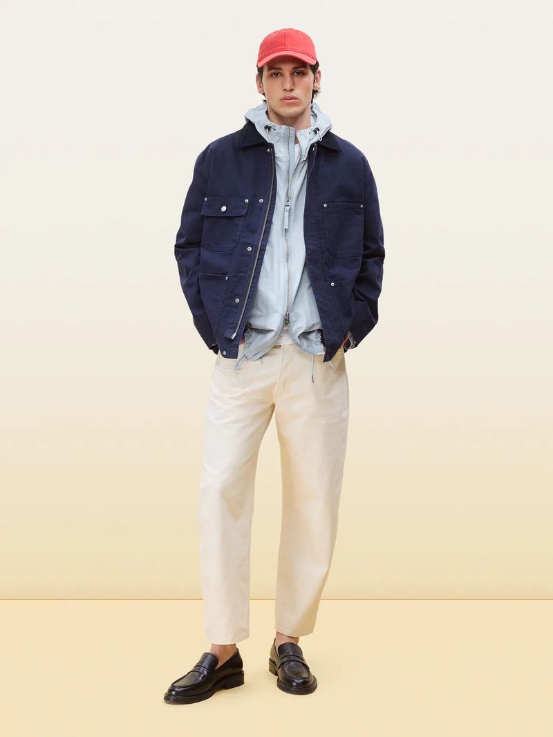 Full-length studio shot of a male fashion model in a navy jacket, light blue top, beige pants, black loafers, and a red baseball cap.