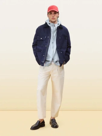 Full-length studio shot of a male fashion model in a navy jacket, light blue top, beige pants, black loafers, and a red baseball cap.