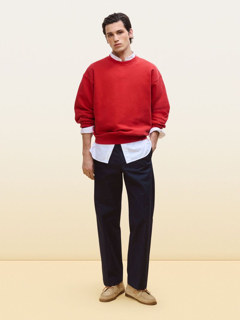 Full body shot of a young adult man in a red sweatshirt and navy pants. He is standing in a studio with a neutral background.