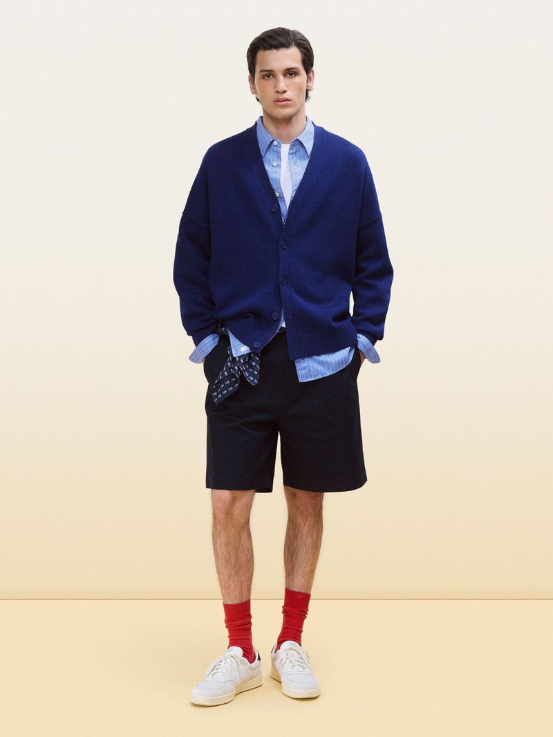 Full shot of a young adult man in a blue cardigan, light blue shirt, dark shorts, and red socks, posing against a light gradient backdrop.