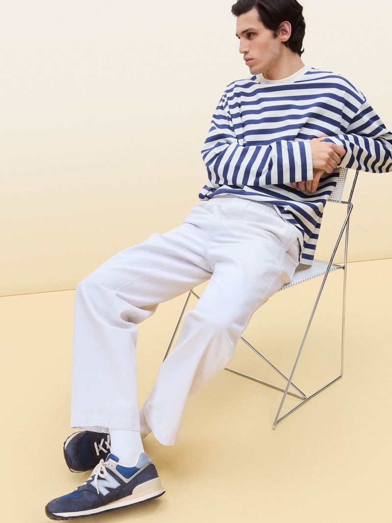 A stylish adult male sits in a wire chair, wearing a blue and white striped long-sleeve shirt and white pants against a light-yellow background.