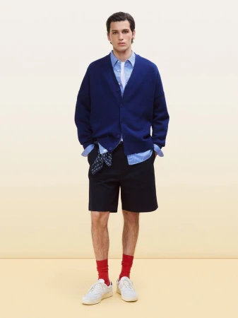 Full-length portrait of a male model wearing a blue cardigan, striped shirt, and dark shorts against a pale background.