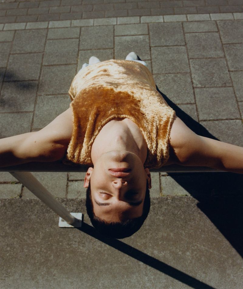 A young adult man lies on a grey tiled surface in a gold, velvet top, relaxing with his eyes closed in the sunlight. Fashion and lifestyle.