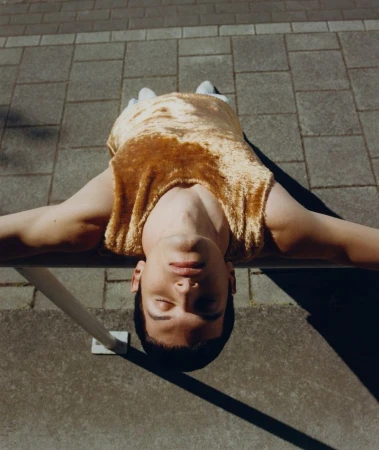 A young adult man lies on a grey tiled surface in a gold, velvet top, relaxing with his eyes closed in the sunlight. Fashion and lifestyle.
