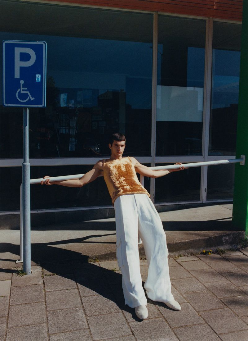 Male fashion model leans against a metal railing in front of a building with windows, wearing a yellow top and baggy white pants.