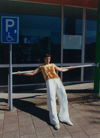 Male fashion model leans against a metal railing in front of a building with windows, wearing a yellow top and baggy white pants.