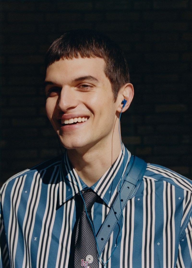 Smiling man in a blue striped shirt and patterned tie poses indoors, wearing wired earphones, with a brick wall visible in the background.