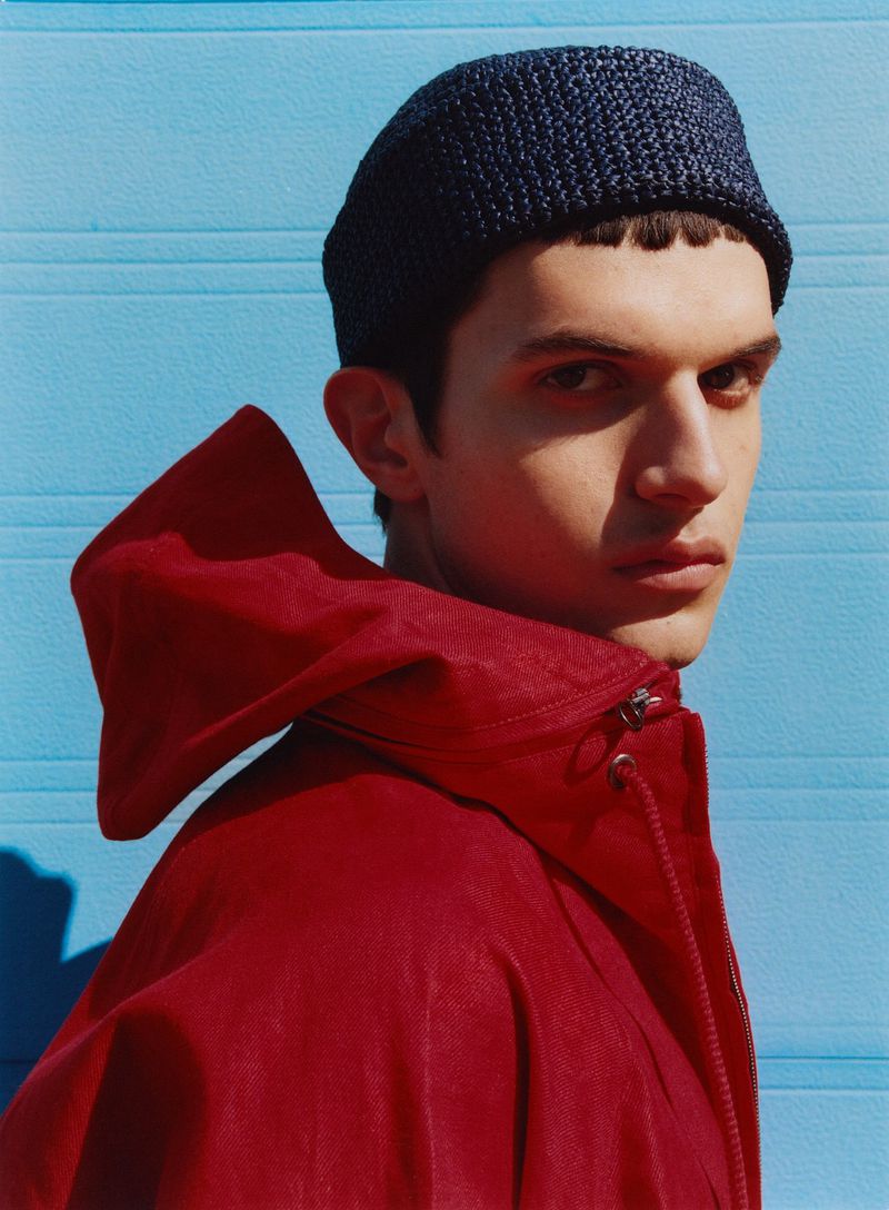 Close-up portrait of a young adult man wearing a knit cap and red hooded jacket against a blue wall. Fashion portrait, studio shot.