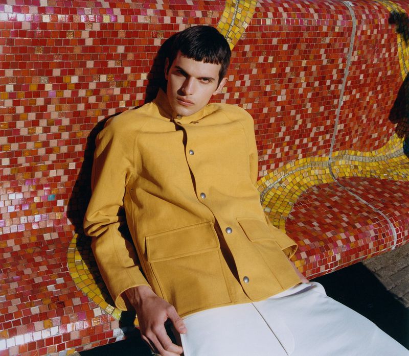 Young adult in a yellow jacket and white pants leans against a mosaic tile wall in an outdoor setting on a sunny day.