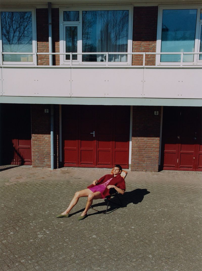 Adult male relaxes in a chair in front of a brick building with red doors, wearing pink shorts and a red shirt in the sun.