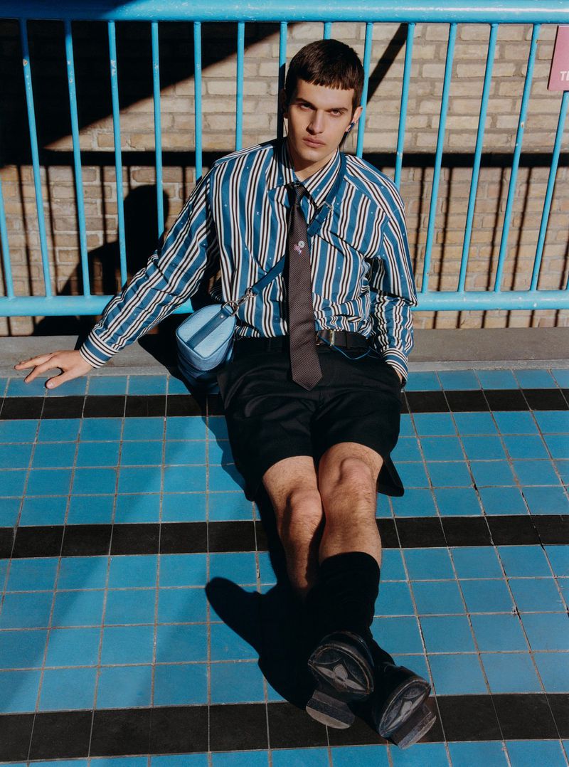 Man in striped shirt and shorts sits on blue-tiled surface with black stripes, posing with a shoulder bag and a relaxed expression.
