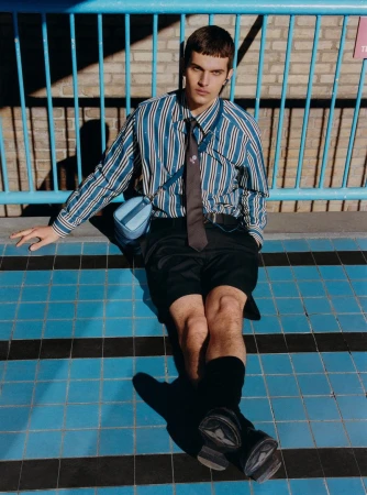 Man in striped shirt and shorts sits on blue-tiled surface with black stripes, posing with a shoulder bag and a relaxed expression.