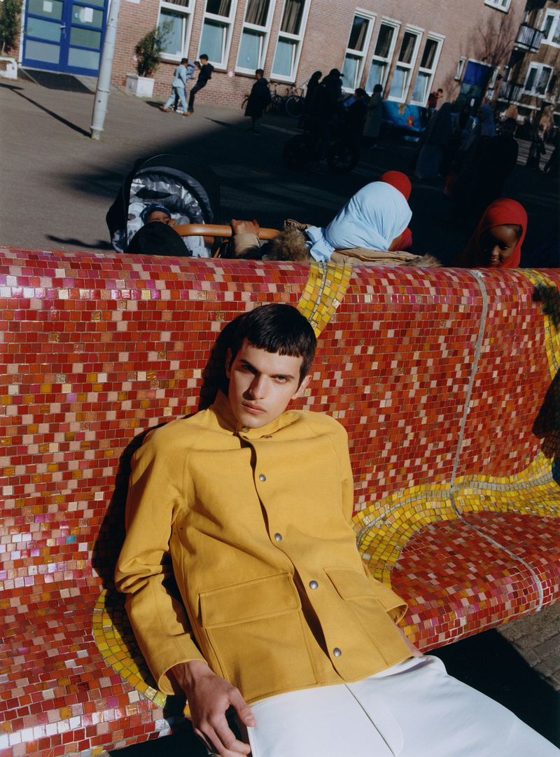Young man in yellow jacket and white pants sits on a colourful mosaic bench on a sunny day in an urban environment.
