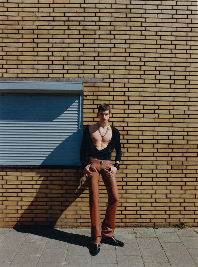 A slender man poses against a brick wall wearing brown leather pants and a black long-sleeved shirt, casting shadows on the textured sidewalk.