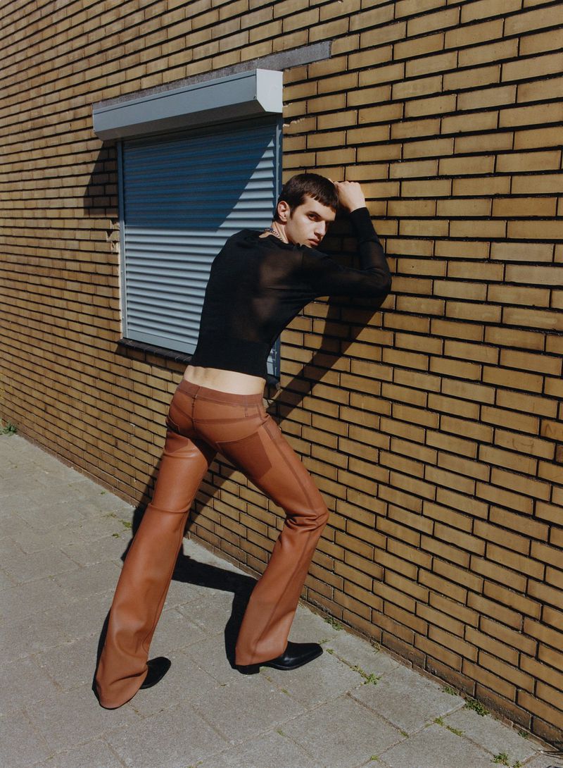 A male model poses outdoors in a black mesh shirt and brown leather pants, leaning against a brick wall with a roll-up garage door.