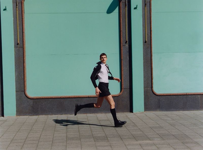 An adult male is running on a sidewalk, dressed in a white shirt, dark shorts, socks, and shoes, against a teal-colored wall outdoors.