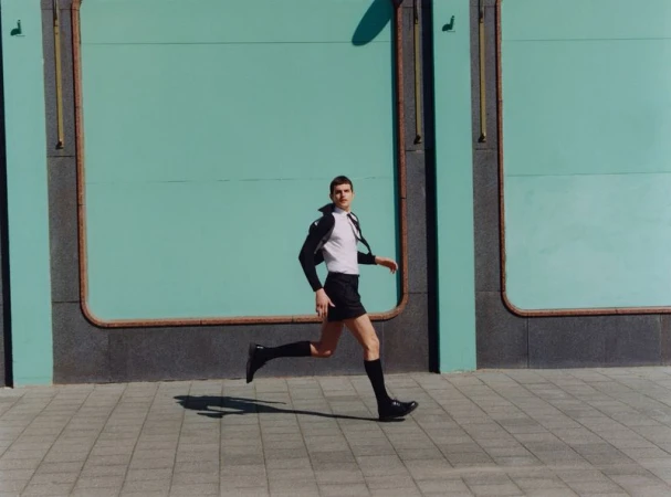 An adult male is running on a sidewalk, dressed in a white shirt, dark shorts, socks, and shoes, against a teal-colored wall outdoors.