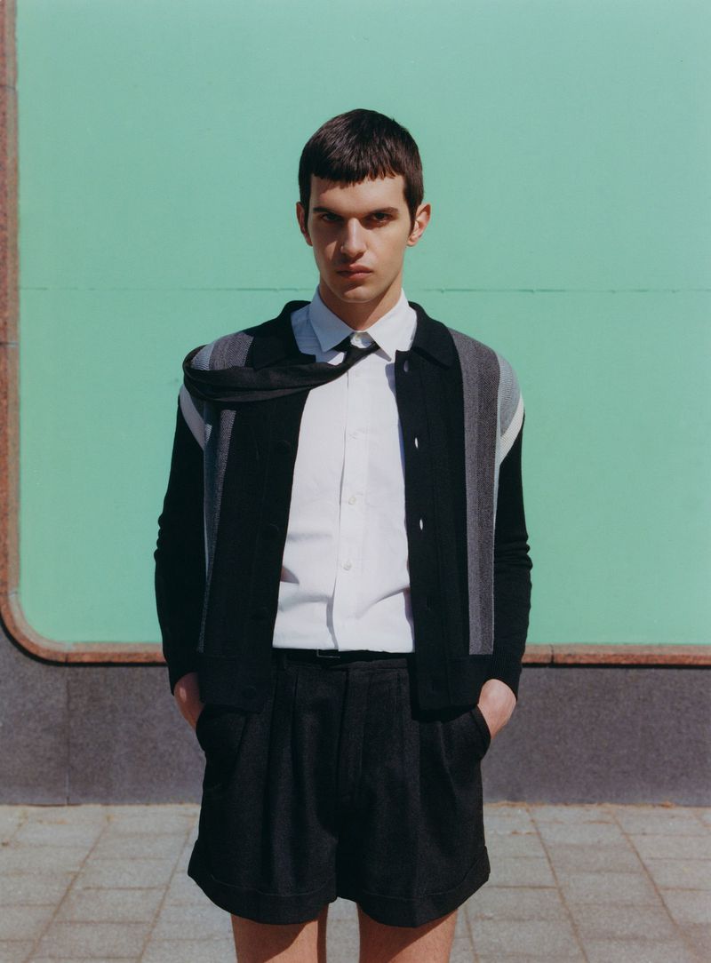 A young man in a black and grey sweater with a white shirt and black shorts stands against a light green background in a medium shot.