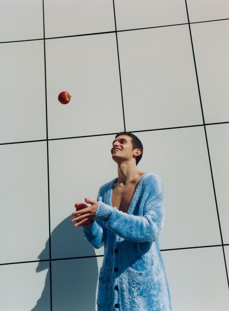 A young man wearing a blue cardigan looking up at a red apple floating in the air with a white wall and black grid background.