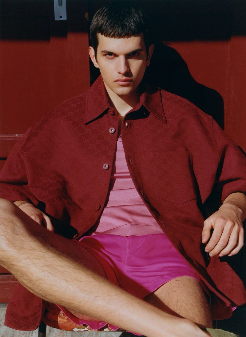 Young male model with dark hair and fair skin poses while sitting against a red metal backdrop, wearing a pink top and red jacket.