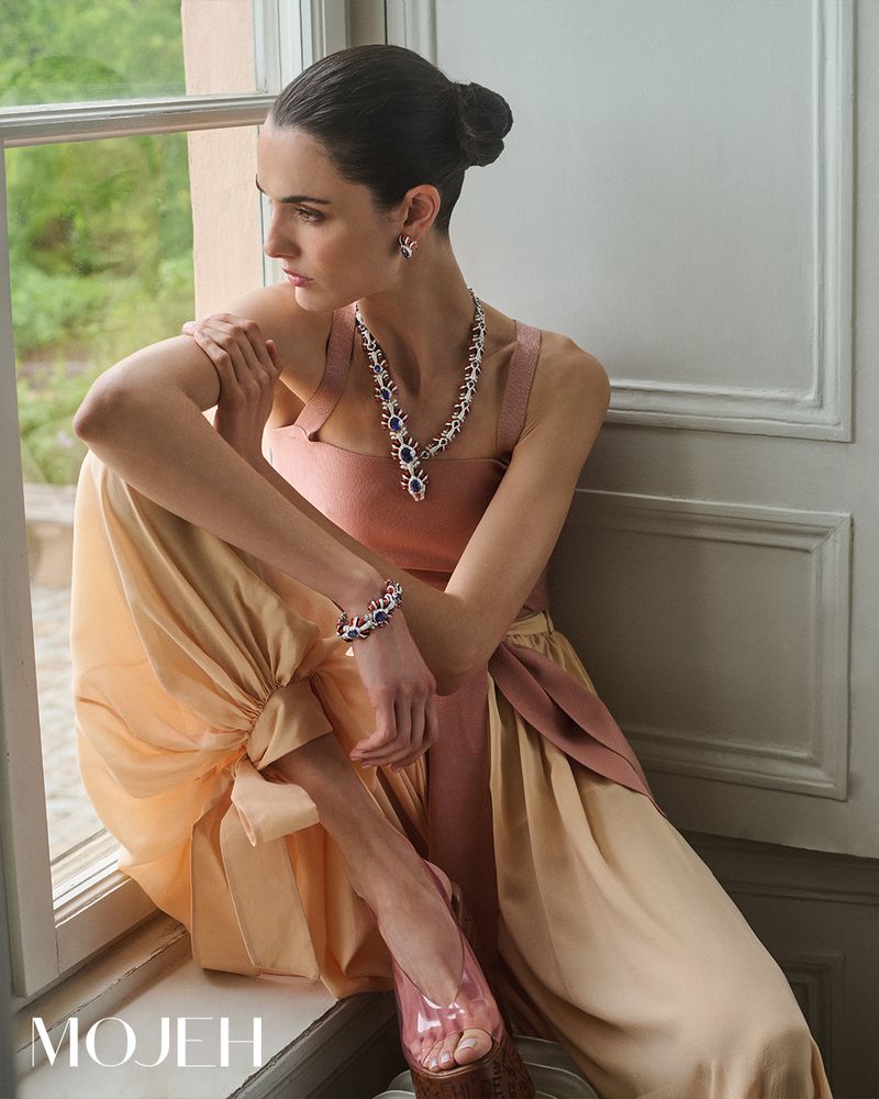 Elegant woman poses in a window with jewelry, wearing a sleeveless top, wide leg pants and platform sandals inside a neutral room.
