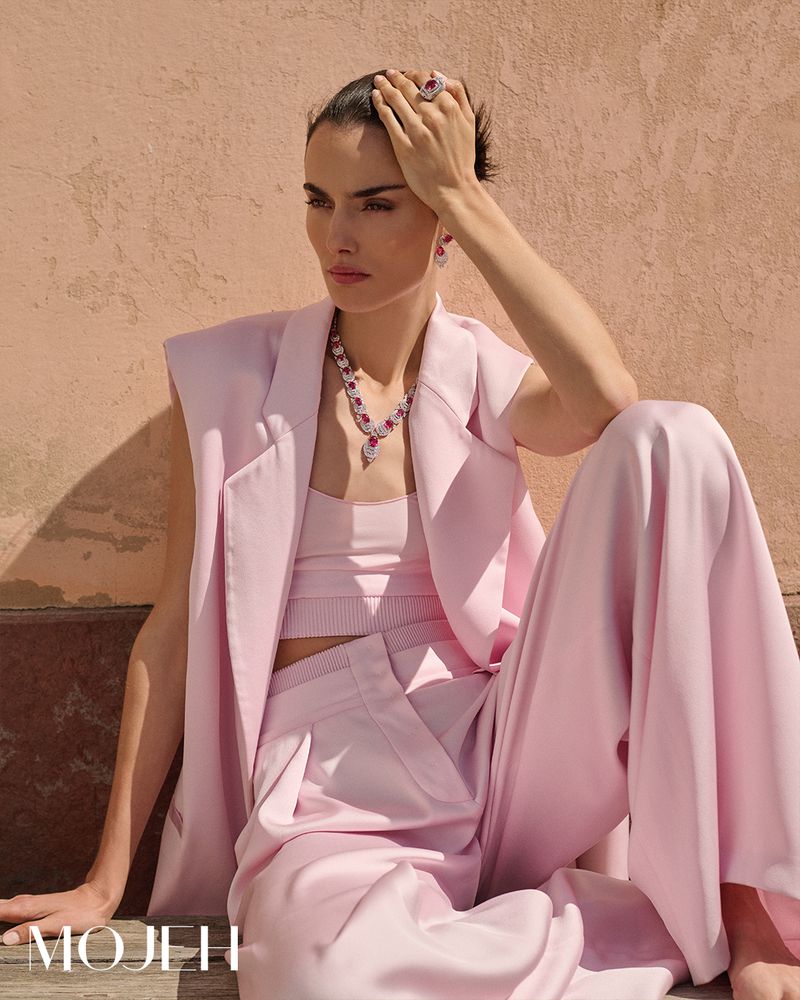 Elegant woman poses in a pink pantsuit and jewelry by MOJH II, showcasing fashion, beauty, and style against a textured wall backdrop.