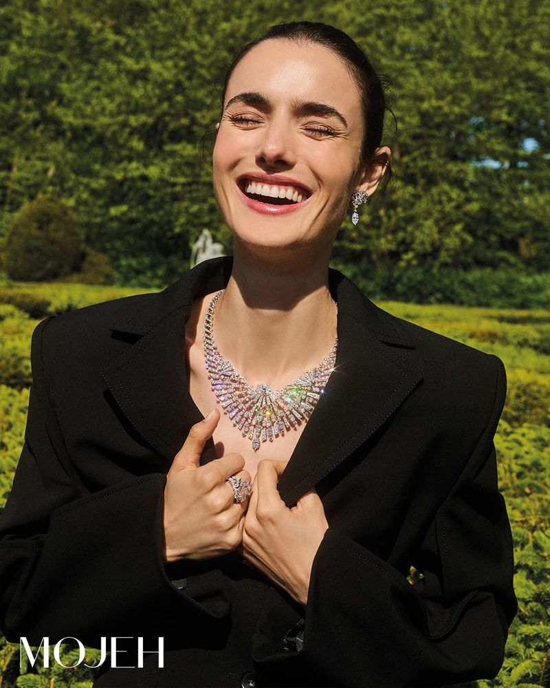 Woman smiles wearing a black blazer and diamond jewelry in a garden setting, capturing a fashion-forward outdoor portrait.