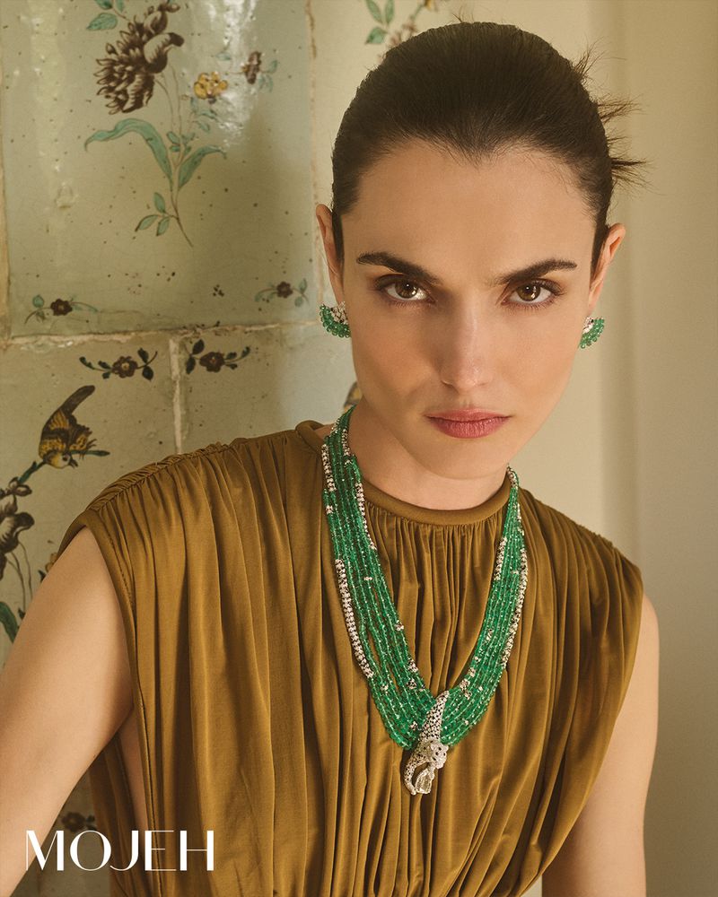 Close-up of a woman with dark hair wearing a sleeveless, draped, gold-brown dress and a tiered green and white beaded necklace and earrings.