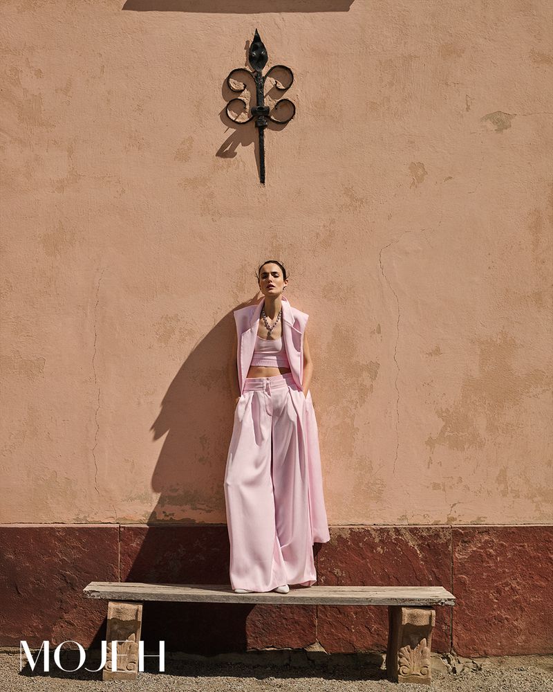 Woman in pink outfit posing in front of a peach colored wall with wrought iron decoration and a stone bench on an outdoor setting.