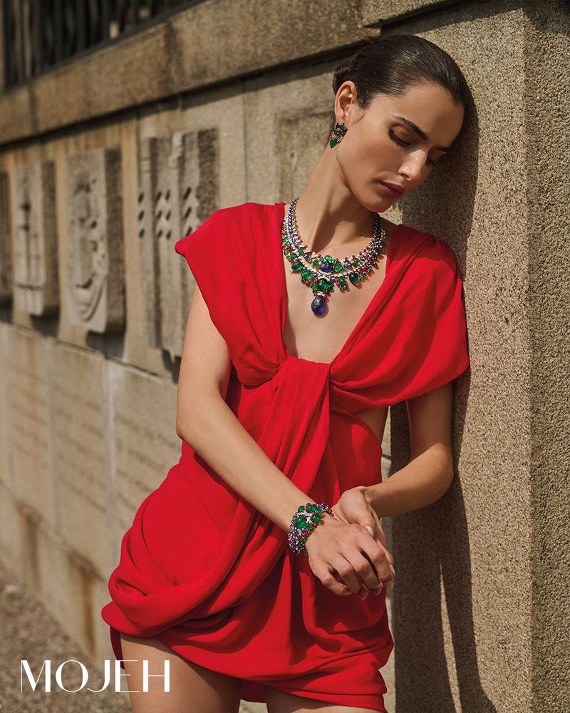 A woman poses in a red dress with emerald jewelry near a concrete wall, showcasing fashion and beauty in an outdoor setting.