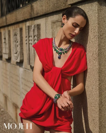 A woman poses in a red dress with emerald jewelry near a concrete wall, showcasing fashion and beauty in an outdoor setting.