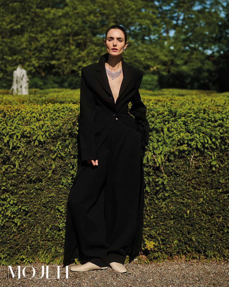 A woman in a black coat and necklace stands in an outdoor garden maze, with lush greenery and a stone fountain in the background.
