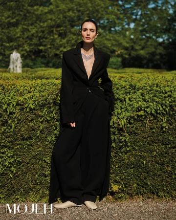 A woman in a black coat and necklace stands in an outdoor garden maze, with lush greenery and a stone fountain in the background.