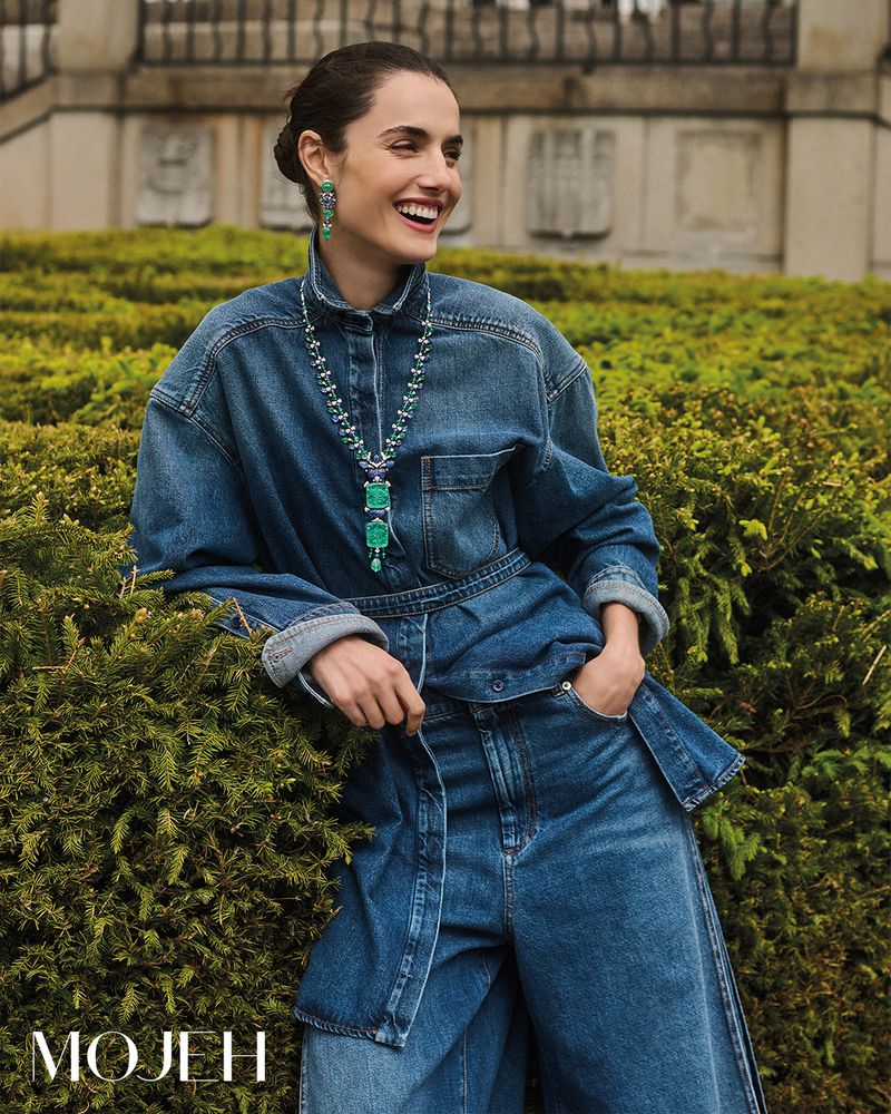Smiling woman in denim shirt and wide leg jeans stands in a garden with shrubbery and an ornate building in the background.