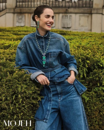 Smiling woman in denim shirt and wide leg jeans stands in a garden with shrubbery and an ornate building in the background.