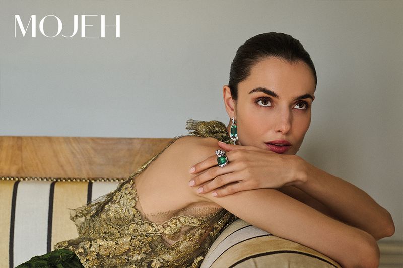 Elegant woman with emerald jewelry reclines on a striped cushion, wearing a gold lace top in a softly lit, neutral setting.