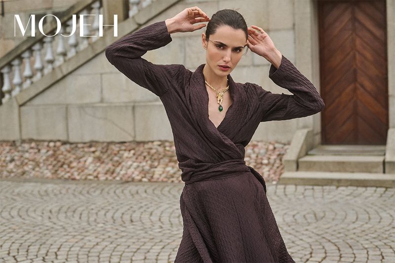 Elegant woman in a wrap dress and statement necklace poses outdoors on stone steps, showcasing fashion and style in neutral tones.