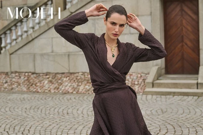 Elegant woman in a wrap dress and statement necklace poses outdoors on stone steps, showcasing fashion and style in neutral tones.