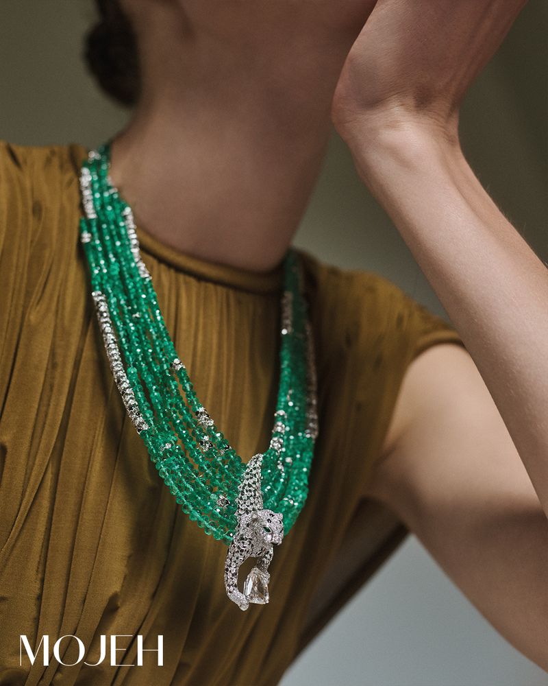 Woman showcases a multi-strand emerald necklace with a jeweled panther pendant, complemented by a mustard-colored top, in a studio setting.