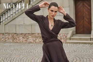 Woman in a dark brown textured wrap dress wearing a gold statement necklace standing outside a stone building