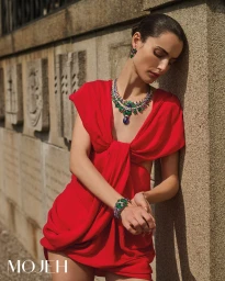 Model in a bright red draped dress leaning against a stone wall, wearing a jewelled necklace and earrings.