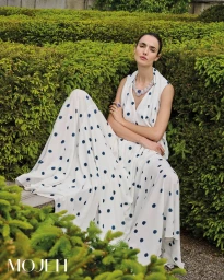 Model in white polka dot dress sitting on a wooden bench amid manicured green hedges for Mojeh Magazine
