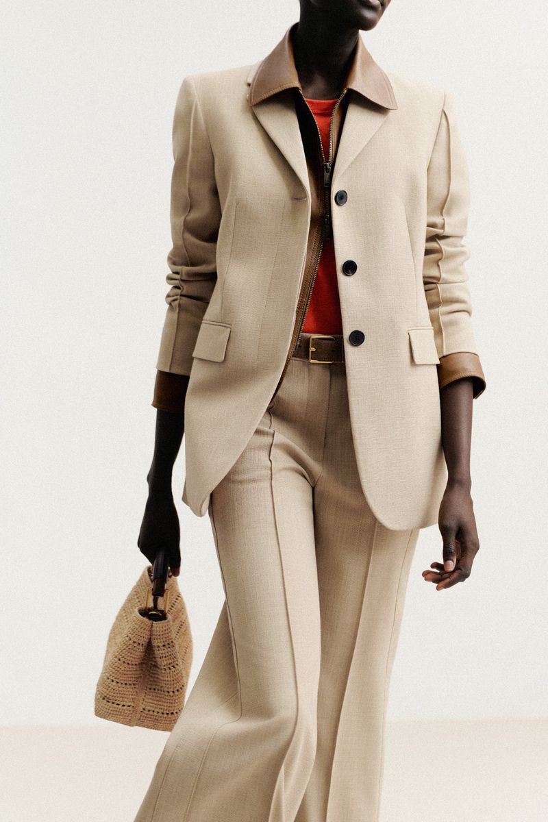 An adult female poses in a tailored neutral-toned suit with wide-leg trousers, complemented by a straw tote bag against a bright, neutral backdrop.