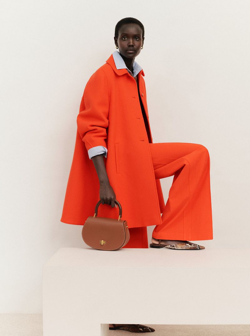 Elegant African woman in vibrant orange coat and trousers poses indoors with a brown leather purse, complemented by a neutral backdrop.